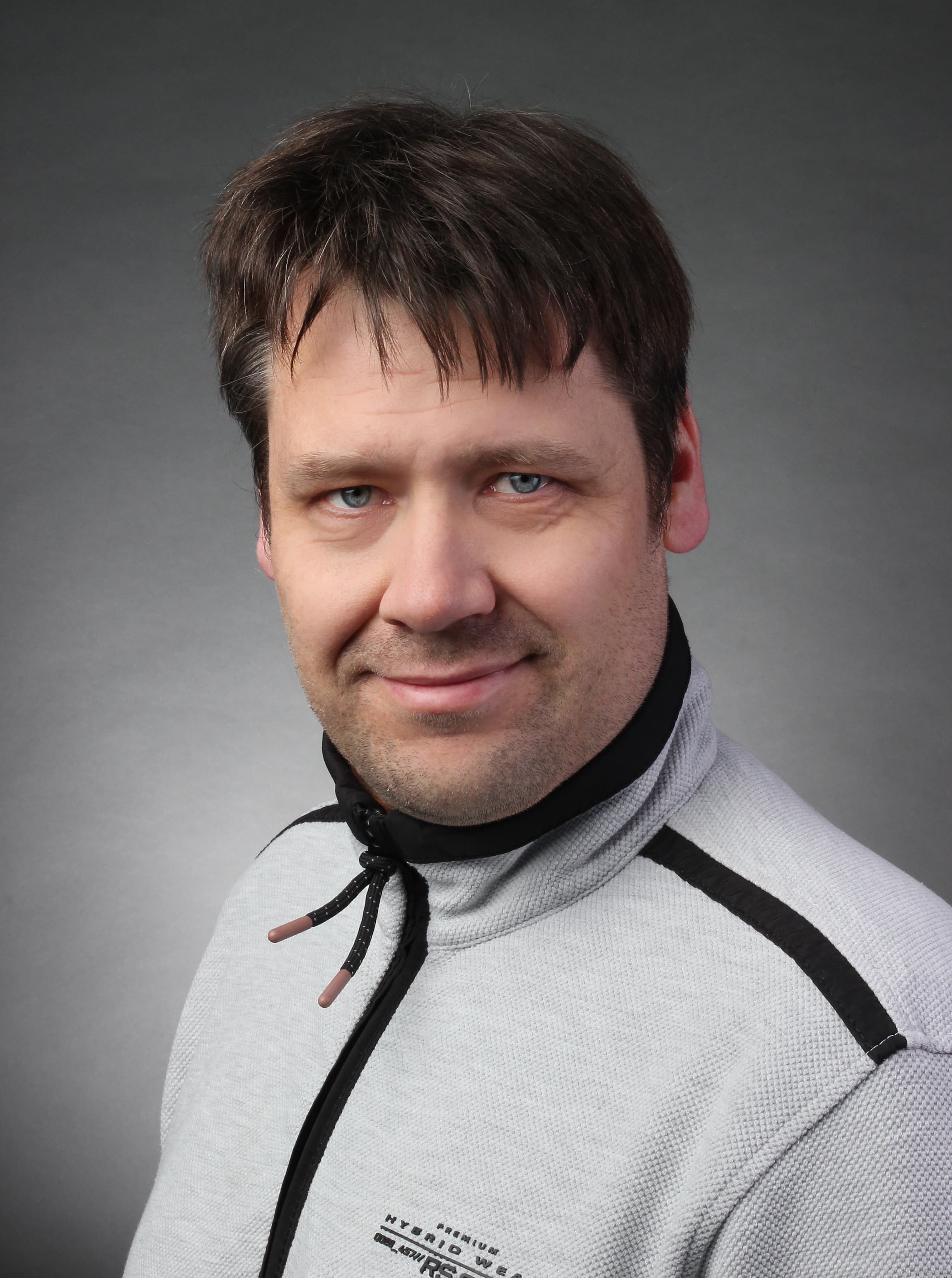 Lächelnder Mann mit braunen Haaren und blauen Augen trägt eine graue Fleecejacke vor grauem Hintergrund.